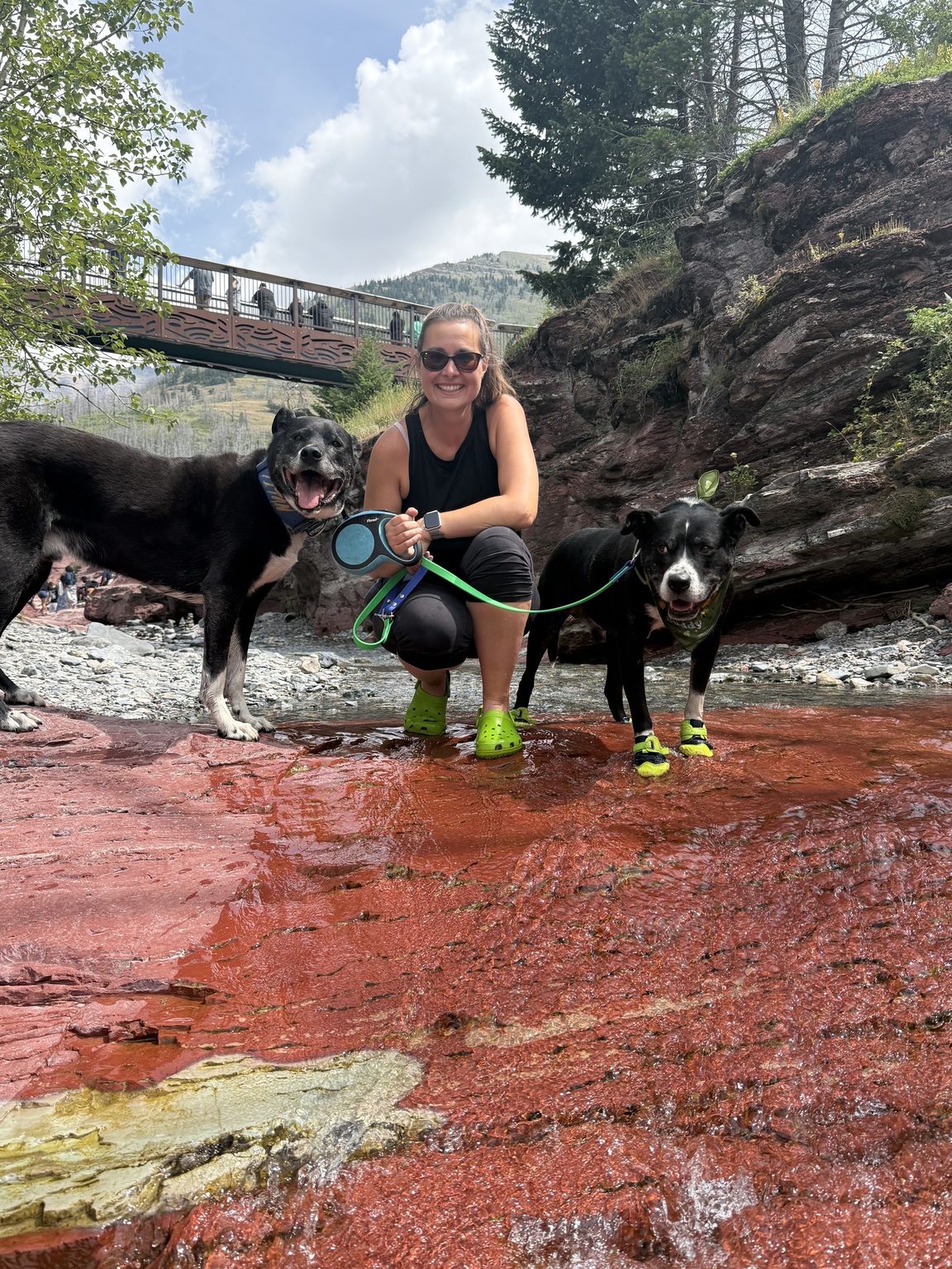 Coreena crouching in a shallow rust-red creek bed with Tuckie and his brother Titan, both wearing water shoes, in Waterton's Red Rock Canyon.