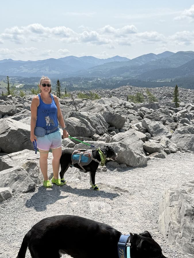 Coreena standing on a rocky mountain trail with Tuckie in a hiking backpack and boots, and Titan in the foreground.